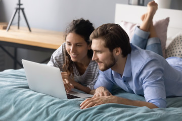 Happy young man and woman relax lying in cozy bed browsing fast wireless internet shopping on laptop together, millennial Caucasian couple rest in bedroom watch video using modern computer at home