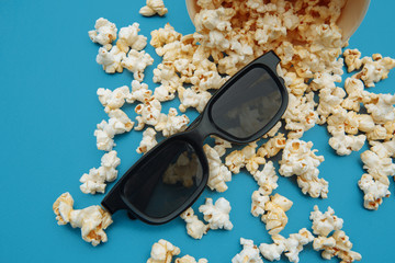 Popcorn spilled out of the bowl. Glasses for viewing the movie are on a blue background.