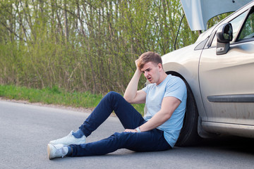 Frustrated sad upset guy driver is sitting near a broken car after road accident, crash. Young...