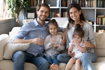 Portrait of happy young Caucasian family with little daughters on couch at home recommend drinking...