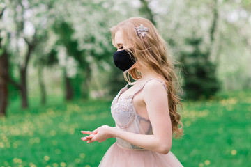 Beautiful young bride in a wedding dress and a black medical mask on her face near a blooming apple trees. Covid-19 protection.