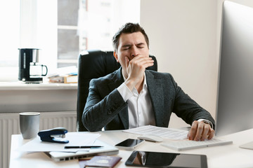 An overworked employee works on a desktop computer. Tired businessman or office worker yawns and falls asleep at the workplace. Sales manager, financial analyst or IT specialist, freelancer.