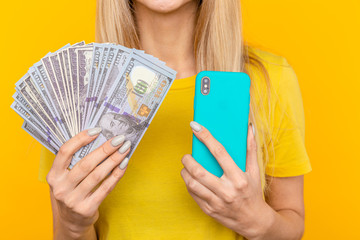 Portrait young girl with long blonde hair standing over yellow background, holding money banknotes, using mobile phone.