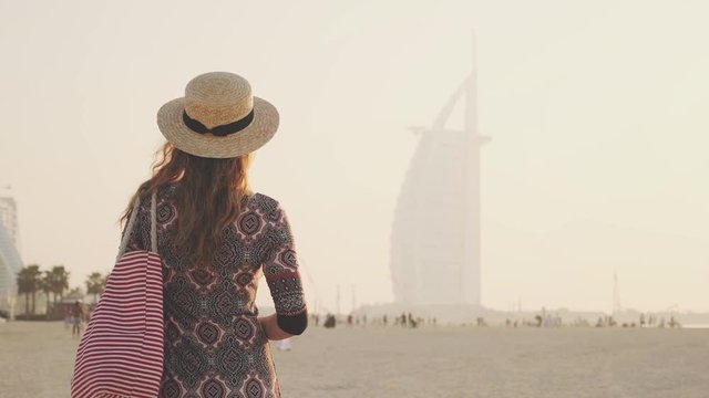 woman tourist enjoy summer vacation on beach coastline sandy plage. Blonde girl turned away looks at blurred simbol Dubai city at foggy sunset. Straw hat striped pink white bag back view. UAE 2020 4k