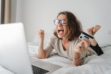 Pretty happy young curly woman with glasses spends time online shopping in bed, with a credit card in her hands with laptop