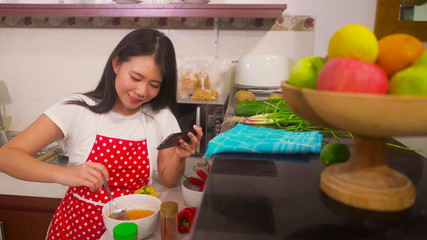 cheerful domestic cooking - young happy and beautiful Asian Korean home cook woman in red apron reading healthy recipe in mobile phone preparing delicious meal in kitchen