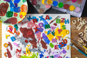 A messy wooden table with colorful watercolor palette, different brushes and letters.