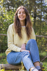 Obraz premium Pretty young brunette woman laughing and looking at camera sitting in a park outdoors on a sunny day. She is wearing blue jeans and a yellow sweater and her hair is down. Vertical image.