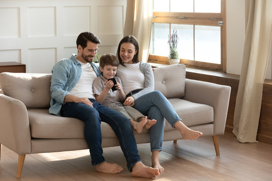 Happy young Caucasian family with son sit relax on sofa in living room using modern smartphone gadget together, smiling parents rest on couch at home with small boy child, browsing cellphone device