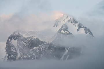 Beautiful alpine scenery with snowy mountains inside low clouds at sunrise. Big glacier in cloudy sky. Morning light through clouds. Scenic minimalist landscape with rocks in dense fog in pastel tones