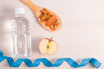 half an apple, a bottle of water, a large wooden spoon with dry apples and a blue measuring tape on a wooden table, proper nutrition concept, maintaining water balance, copy spcae, top view