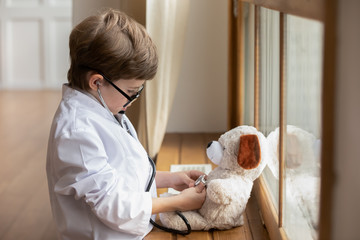 Caring little boy kid in whites and glasses listen to plush toy heartbeat with stethoscope, cute small child play act as doctor cure examine teddy bear heart in hospital, healthcare, medicine concept - obrazy, fototapety, plakaty