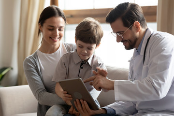 Positive male pediatrician sit on couch with little boy patient with mom show anamnesis on tablet, caring man doctor with small child and mom client use pad gadget for lab results in modern hospital - obrazy, fototapety, plakaty