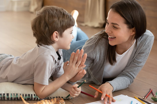 Overjoyed young mother and small preschooler son give high five playing drawing together, happy mom or nanny have fun painting on paper learning with little boy child at home, education concept