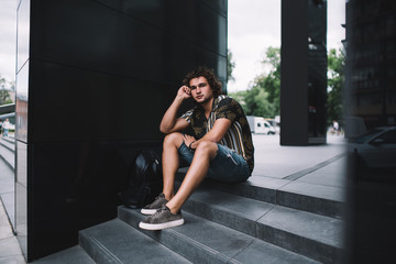 Fototapeta na wymiar millennial hipster guy traveler looking at camera having rest on street in city dressed in trendy wear, handsome male tourist in stylish casual outfit sitting on urban setting background with bag