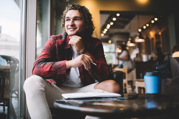 Positive blonde male student in casual wear enjoying learning process in coffee shop looking at window,smiling man blogger using notepad for creating article publication spending time in cafe interior