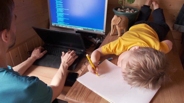 father work at home on computer, small son child lie near and paint in album, concept of stay home in pandemic or quarantine, self-isolated family candid lifestyle, parenting