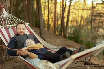 Young family hipster playing with son on a hammock in park on camping trip, family vacation