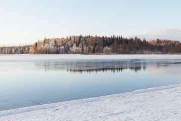 Unfrozen lake with snowy coastline with a forest at background.