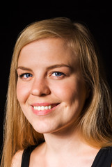 Portrait of a beautiful natural blonde with blue eyes, on a black background. She looks at the camera, smiling a confidential smile.