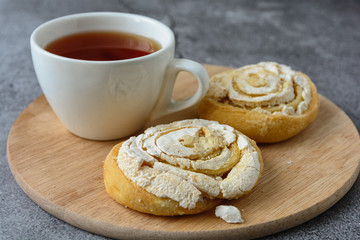 Two cakes in the form of a snail with curd filling, topped with sugar icing, is on a wooden round board next to a cup of tea