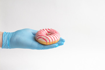 Person in blue surgical gloves holds striped donut on his hand on white background. Theme of Safe food delivery during coronavirus pandemic.
