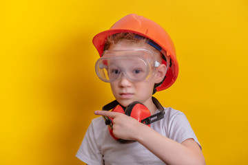 Cute boy in builder glasses, helmet and headphone to ear protection shown finger up over yellow background  copy space