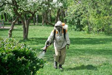 Fototapeta premium A man in a protective suit conducts anti-mite treatment in a city park. Spraying repellent among green grass and blooming apple trees. Repelling and killing insects
