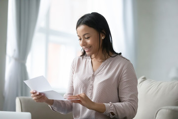 Young African woman sitting on sofa in living room reading postal letter feels happy. Getting bank...