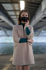 Elegant woman under a bridge wears a black medical mask to protect herself from coronavirus COVID-19, urban portrait in Russia. 