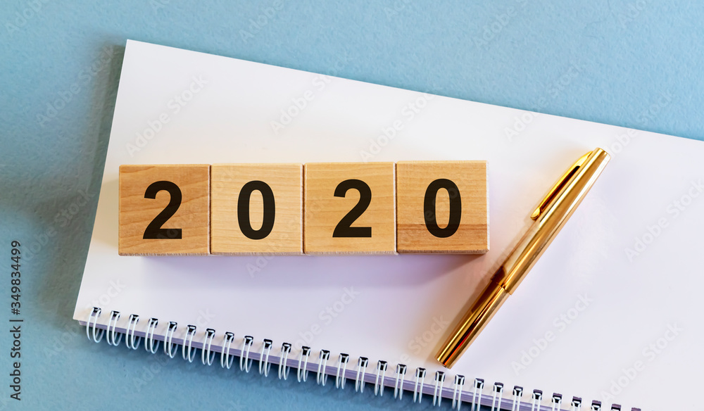 Poster year 2020 sign text number on wooden cube blocks on a white notebook planner and golden pen on blue 