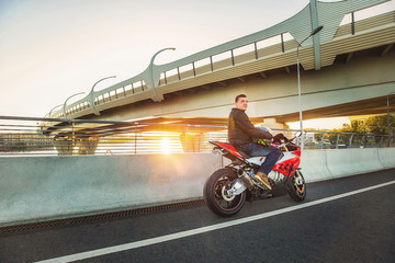 Handsome young man wearing casual outfit sitting on red sports motorcycle on the road in sunset with helmet in hand