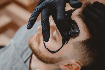Naklejka premium Acne removal with cosmetic product, cleaning pores of men skin, barber applying black charcoal mask to client man in spa salon