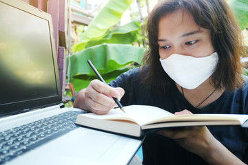 Smile Asian Thai woman wearing a white cloth mask for prevent the Covid-19 or Corona virus and writing for work from home with laptop and working online system. Air Pollution Value Pm 2.5 concept.