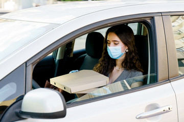 Lovely caucasian girl with green eyes looks at pizza and wants to eat. Young girl holds pizza in car and works as courier in delivery company at quarantine covid 19. Courier in medical mask and gloves