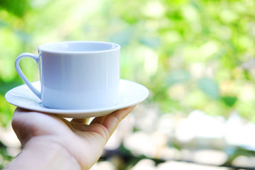 Hand holding white ceramic cup of hot coffee with natural light morning in the garden. Caffeine drinks cause a fresh feeling.