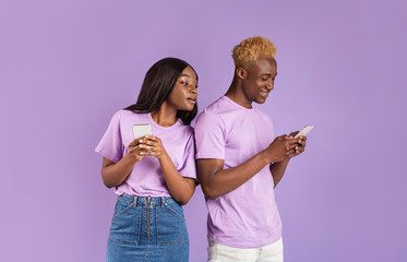 Jealous black girl looking over her boyfriend's shoulder into his smartphone, watching his message on lilac background