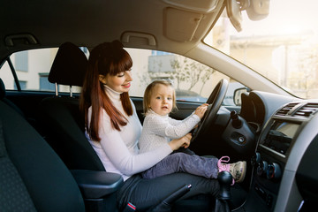 Young mother with her little baby girl sitting in the car in the front seat near car steering wheel. A woman and baby girl pretending to be driving. Mother shows her baby the car inside