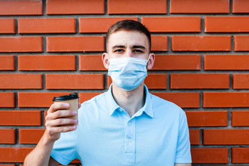 Portrait of a guy in a protective mask on a brick background. Image of a young man's face in a mask to prevent germs. A young man holds coffee in one hand and drinks it through a mask. Coronavirus.