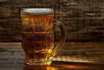 A glass of amber beer. Glasses on a wooden table. Black shabby boards on the background. Bright contrasting light and glare on the glass. Place for text near the booze.