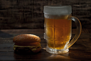 Cheeseburger with a glass of beer on a dark wooden table. Food on a black shabby background. Contrasting yellow light sparkles on the glass. Frozen mug with amber beer.