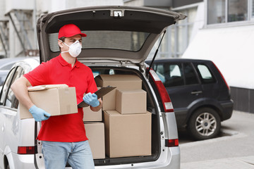 Courier in uniform, protective mask and gloves stands near trunk of car with parcels