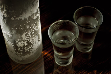 Frozen glasses with alcohol near the bottle. Drinks on a dark brown table. Transparent utensils on a black background. Old shabby boards under glass.