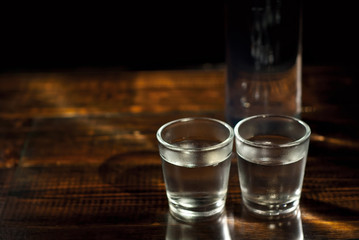 Frozen glasses with alcohol near the bottle. Drinks on a dark brown table. Transparent utensils on a black background. Old shabby boards under glass.