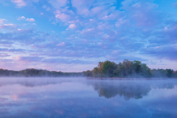 Obraz premium Landscape at dawn of the foggy, spring shoreline of Whitford Lake with reflections in calm water, Fort Custer State Park, Michigan, USA
