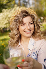Fototapeta premium Women clinking glasses with tasty wine on light background at summer day. Happy blonde with curly hair in a straw hat