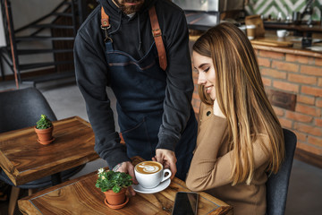 Barista male in blue apron giving order, latte with drawing for blonde woman customer in coffee shop, service mind concept