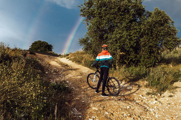 Young cyclist on a country road in the afternoon with the rainbow in the background. Selective focus.
