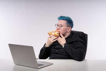 Young man with blue hair is having a snack in front of a laptop, biting a slice of pizza with appetite, studio shot