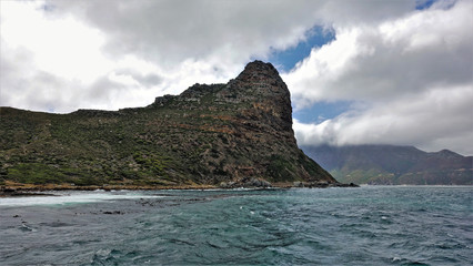 Severe cliffs in the cold atlantic ocean. Steep rocky slopes are covered with sparse shrubs. The waves beat against the shore. Dense picturesque clouds lie on the tops of the mountains. South Africa.